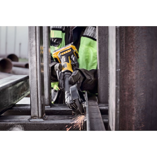 A person wearing protective gloves and a high-visibility jacket uses a DEWALT cordless angle grinder to cut or grind a metal beam, with sparks flying during the process.