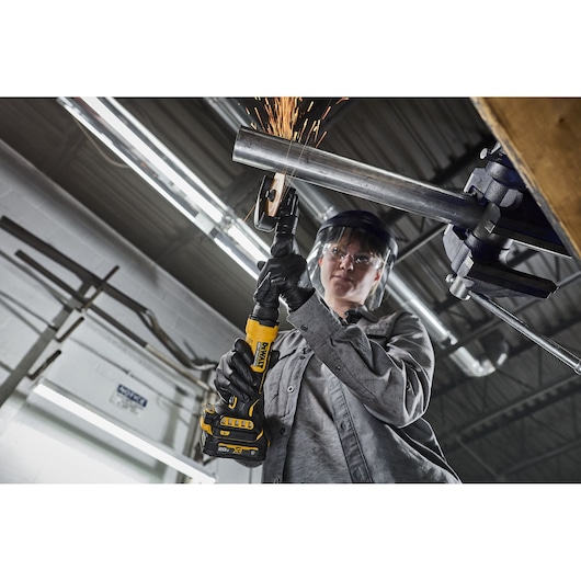 A person wearing safety gear uses a DEWALT cordless power tool to grind a metal pipe, creating sparks. The scene is set in an industrial workshop with overhead lighting and other equipment visible.