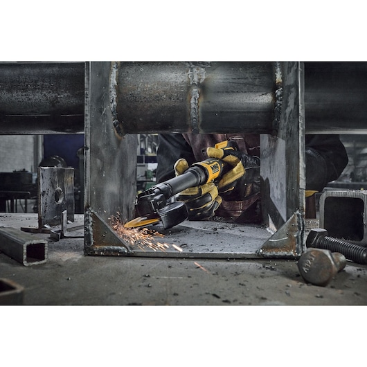 A person wearing gloves uses a DEWALT angle grinder to cut or grind a metal structure, with sparks flying from the contact point. Industrial bolts and metal parts are visible on the worktable.