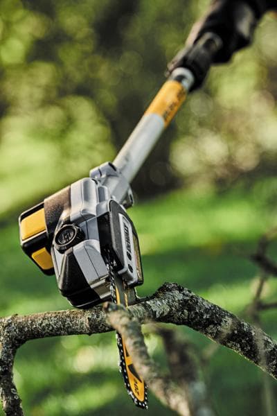 A close-up of a DEWALT pole saw cutting a tree branch outdoors, showing the saw blade slicing through the branch with a blurred green background.