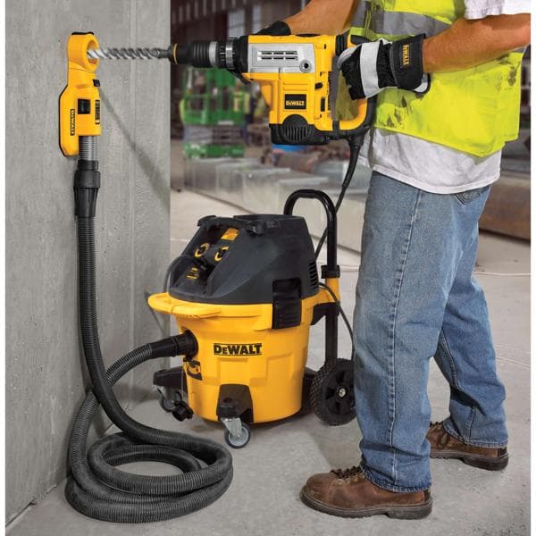 A construction worker uses a DEWALT drill and dust extraction system to drill into a concrete wall. The setup includes a DEWALT vacuum connected by a hose to the drilling area, minimizing dust during operation.