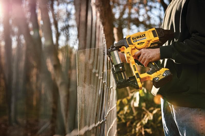 A person using a DEWALT cordless fencing stapler to secure wire fencing outdoors, with trees and sunlight in the background.
