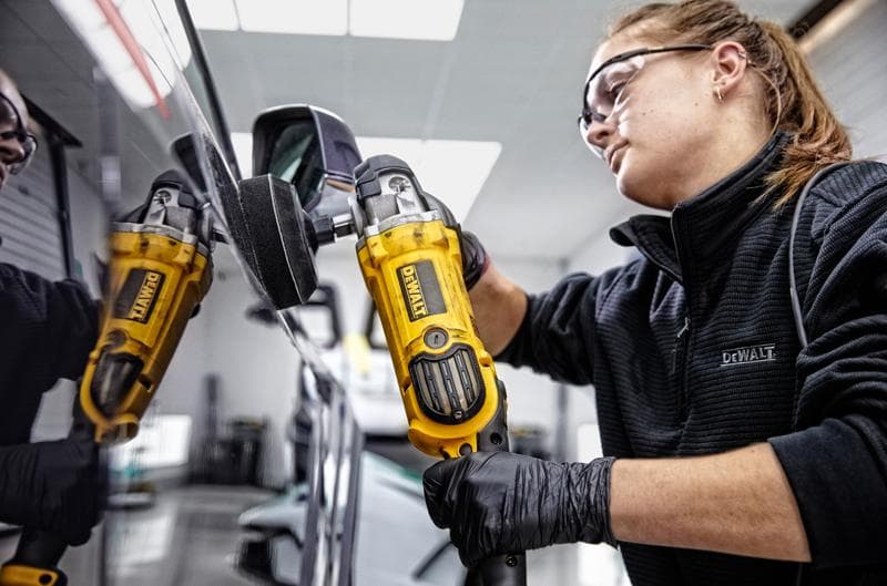 Person uses a yellow DEWALT polisher, model DWP849X, to polish a car side mirror indoors. Face is blurred.