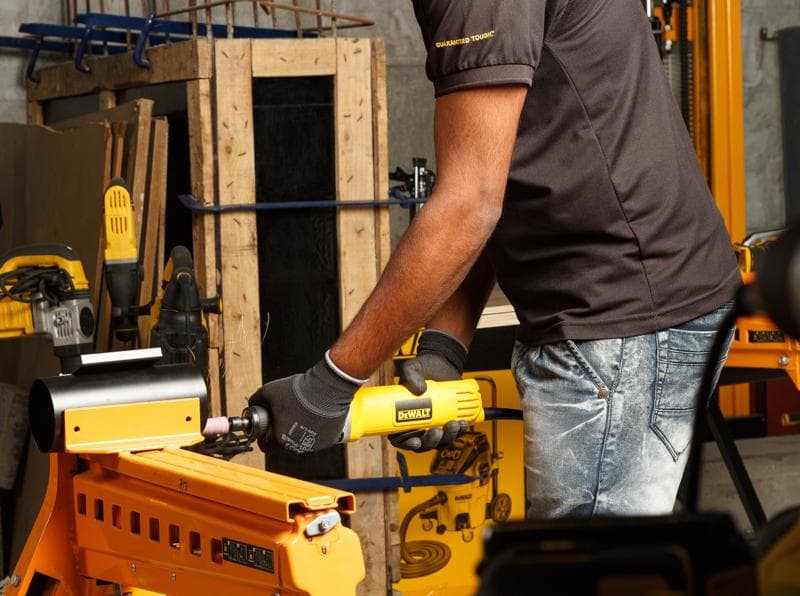 Person using a DEWALT DWE4887N die grinder on metal in a workshop with other yellow tools visible.