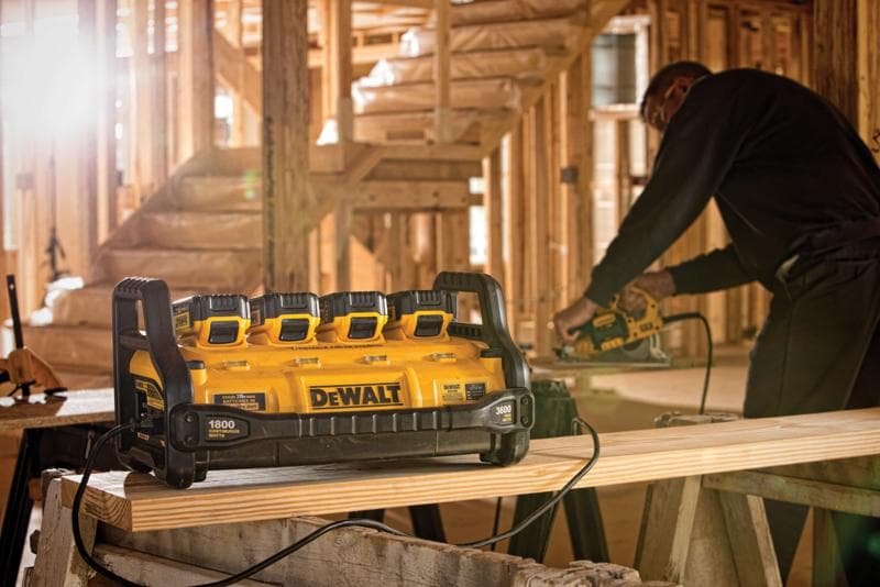 A DEWALT portable power station with four battery slots is shown on a workbench at a construction site. A person in the background is using a power tool on a wooden plank.
