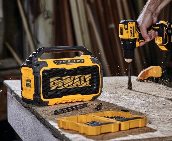 A DEWALT portable Bluetooth speaker positioned on a workbench next to a DEWALT cordless drill in use, drill bits, and a yellow organizer case, all in a workshop setting.
