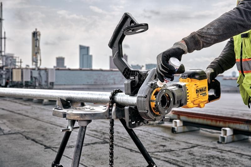 A person using a DEWALT DCE700X2 yellow power tool to thread a metal pipe mounted on a work stand, outdoors on a rooftop with city buildings visible in the background. The user is wearing gloves and a safety vest.