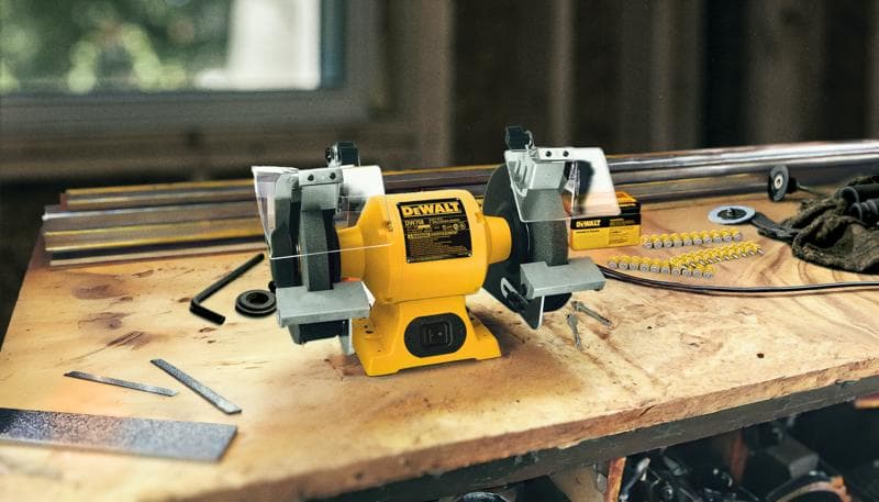 A DEWALT bench grinder, model DW758, is placed on a wooden workbench. Surrounding it are various metal tools and parts, suggesting an active workspace. The grinder is yellow and features two grinding wheels with safety guards.