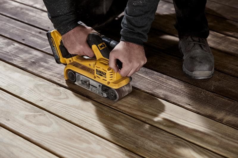 A person using a yellow DEWALT cordless belt sander to sand wooden planks on a floor.