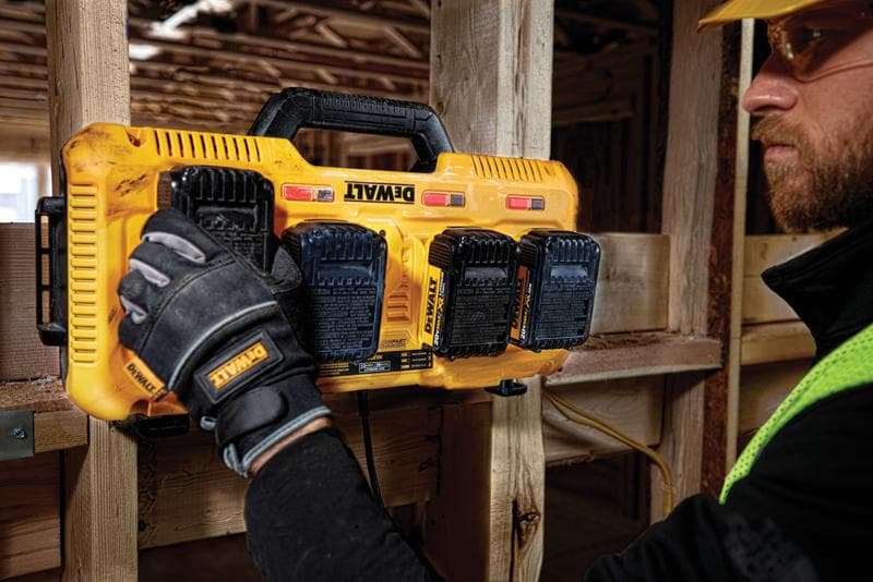 A person wearing work gloves installs or removes batteries from a yellow DEWALT multi-battery charger mounted on a wooden beam at a construction site.