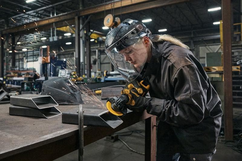 A person wearing protective gear uses a DEWALT DCG400 angle grinder to cut or grind metal pieces on a workbench in an industrial workshop. Sparks are visible, and the environment features various tools and equipment.