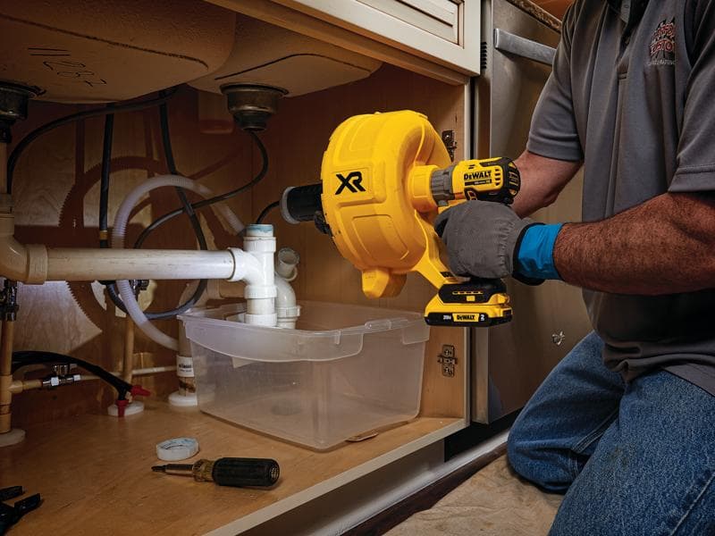 Person using a yellow DEWALT DCD200D1 cordless drain snake tool to unclog a pipe under a kitchen sink. The tool is operated inside a cabinet, with a plastic container placed under the pipes and various plumbing tools nearby.