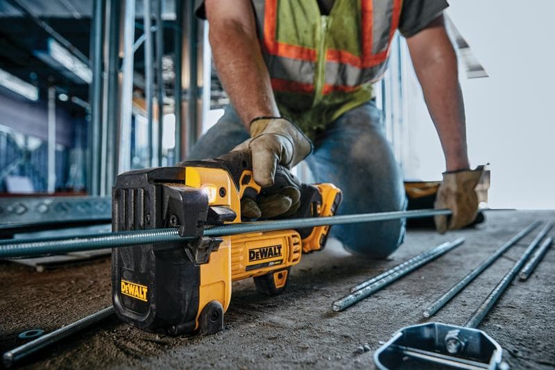 A person wearing work gloves and a safety vest uses a DEWALT cordless cutting tool to cut a metal rod at a construction site.