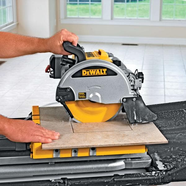 A close-up view of a DEWALT tile saw in use, cutting a ceramic tile on a yellow workstation. Water is visible around the cutting area and the operator's hands are guiding the tile. The saw is being used in a bright tiled room with large windows.