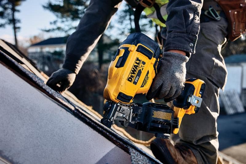 A person wearing work gloves uses a yellow DEWALT cordless roofing nailer on a sloped roof outdoors.