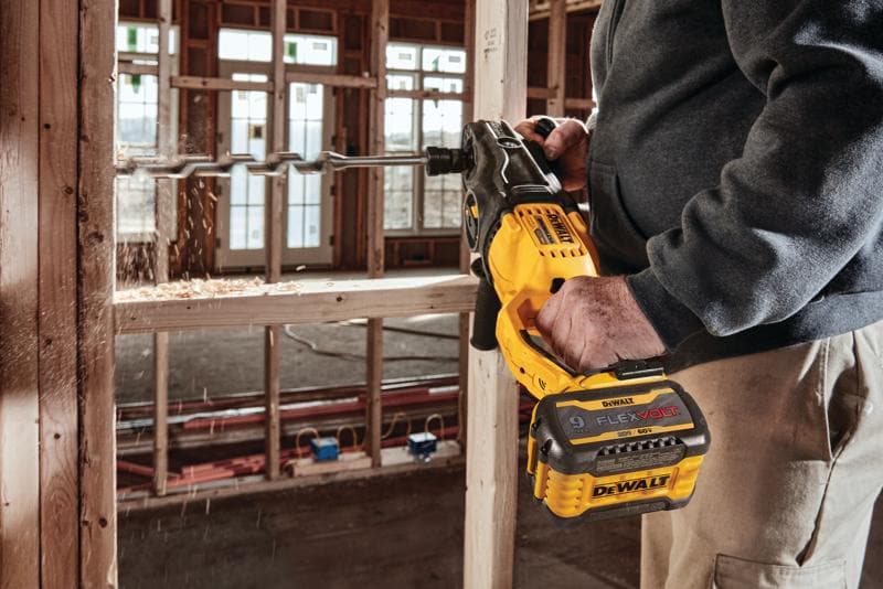 A person is using a DEWALT FlexVolt cordless drill with a large auger bit to drill into a wooden frame at a construction site.