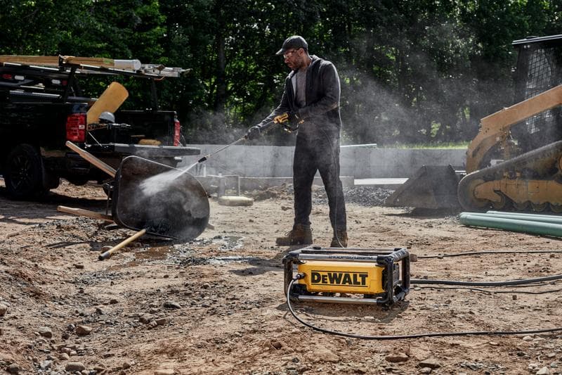 A person using a DEWALT pressure washer to clean a wheelbarrow at a construction site, with construction vehicles and equipment visible in the background.