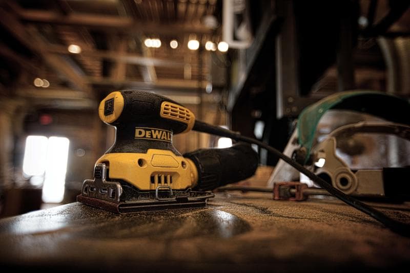 A DEWALT DWE6411K electric sander sits on a dusty workbench in a workshop, with a cord attached and safety equipment visible in the background.