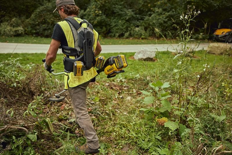 A person wearing a DEWALT battery harness system and a safety vest is working outdoors near a road, using a power tool to clear vegetation. Several DEWALT batteries are attached to the harness for easy access.