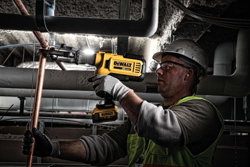 A person wearing safety gear uses a yellow DEWALT cordless tool to cut or shape a copper pipe in an industrial setting.