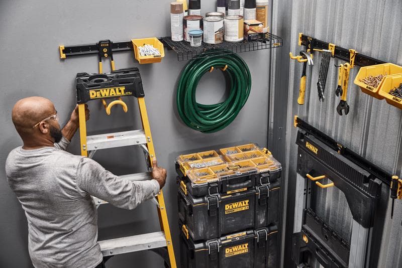 A person organizing a workspace with DEWALT storage products, including a ladder, toolboxes, wall-mounted bins, hand tools, a garden hose, and shelves with paint cans.