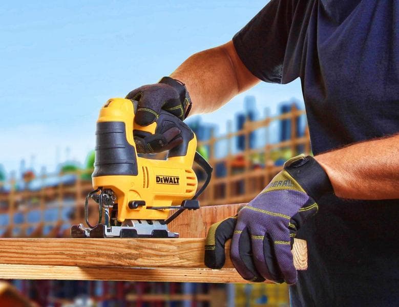 A person wearing protective gloves is using a yellow DEWALT jigsaw to cut a piece of wood, with a construction site visible in the background.