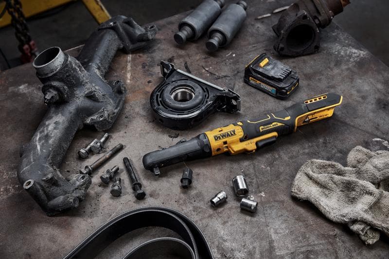 DEWALT DCF510B cordless ratchet tool placed on a workbench, surrounded by various automotive parts, bolts, sockets, battery pack, and a dirty cloth, suggesting a mechanical or repair setting.