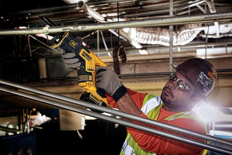 A person wearing protective gloves, a safety vest, and a hard hat is using a DEWALT DCS367B cordless reciprocating saw to cut a metal pipe in an industrial setting. The person's face is intentionally blurred for privacy.