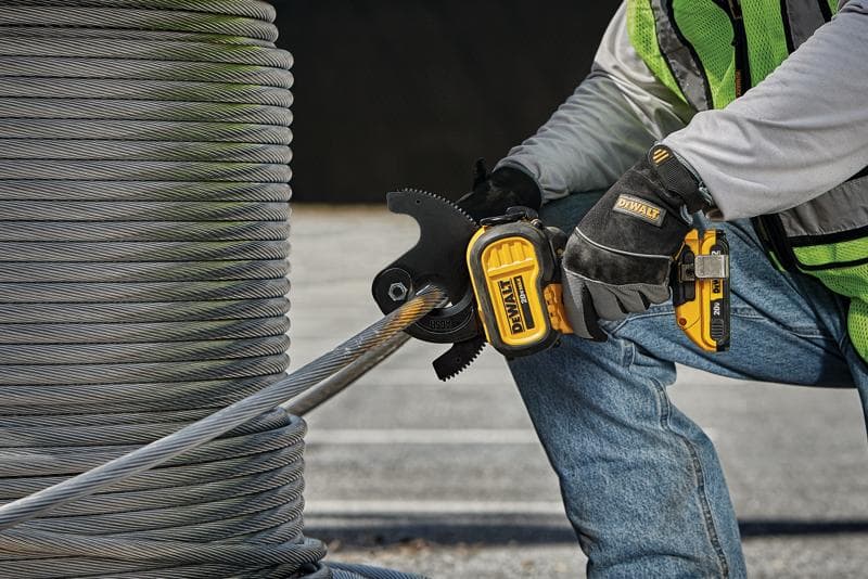 A person wearing safety gloves and a safety vest uses a DEWALT cordless power tool to cut through a thick metal cable next to a large spool of wire outdoors.