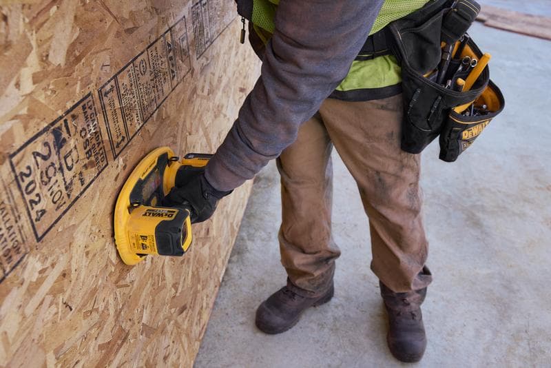 A worker wearing brown pants and protective gloves uses a DEWALT handheld tool device against a large sheet of oriented strand board (OSB) on a construction site. The worker has a tool belt with various hand tools.