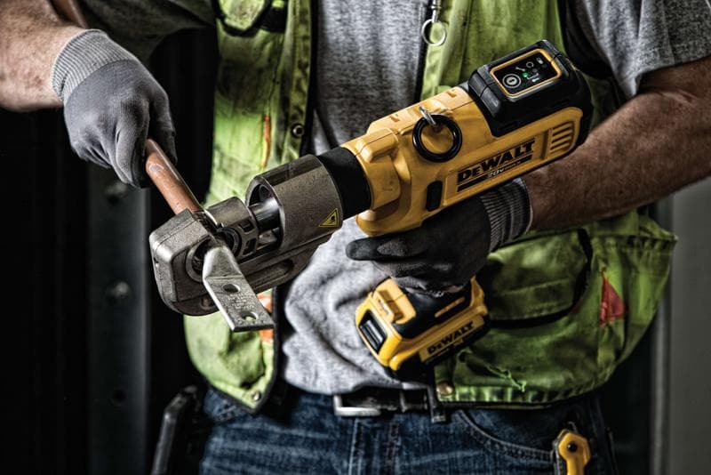 A person using a DEWALT cordless tool to cut a thick metal rod while wearing work gloves and a green safety vest.