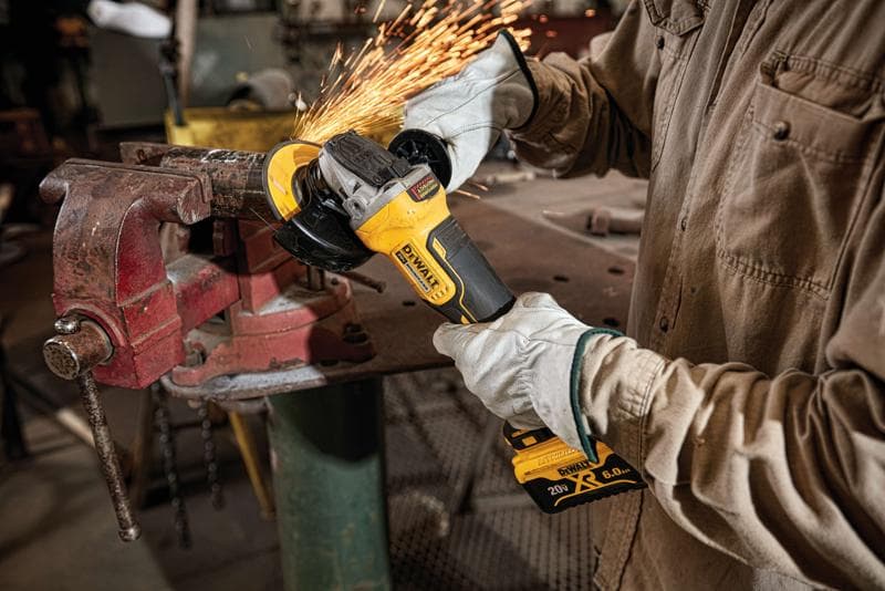 Person wearing protective gloves using a DEWALT cordless angle grinder, model DCG413R2, to cut or grind metal clamped in a vise, with sparks flying in a workshop.
