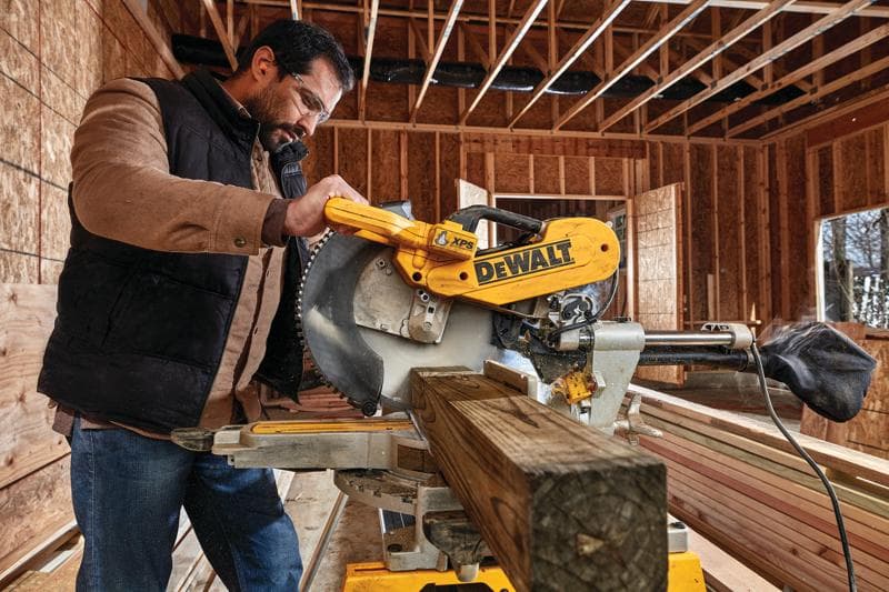A person operates a DEWALT DWS780 miter saw to cut a wooden beam inside a construction site. The saw is yellow and features the DEWALT logo prominently. The background shows exposed wooden framing and unfinished walls.