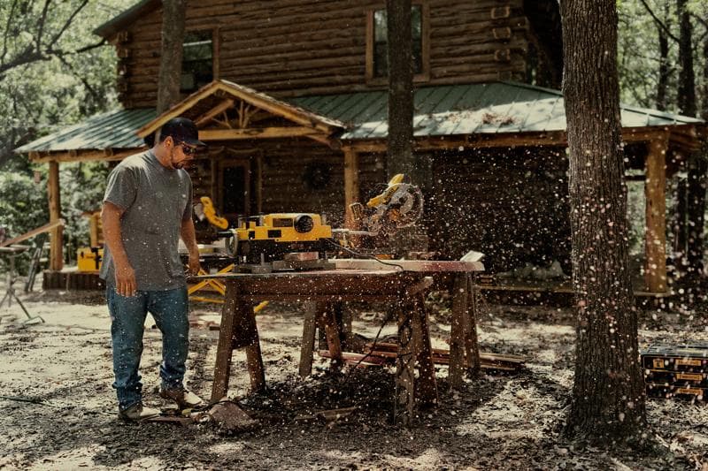 A person using a DEWALT DWS780 miter saw outdoors, cutting wood on a worktable in front of a log cabin. Sawdust is flying in the air as the person operates the tool. Surrounding area includes trees and woodworking equipment.
