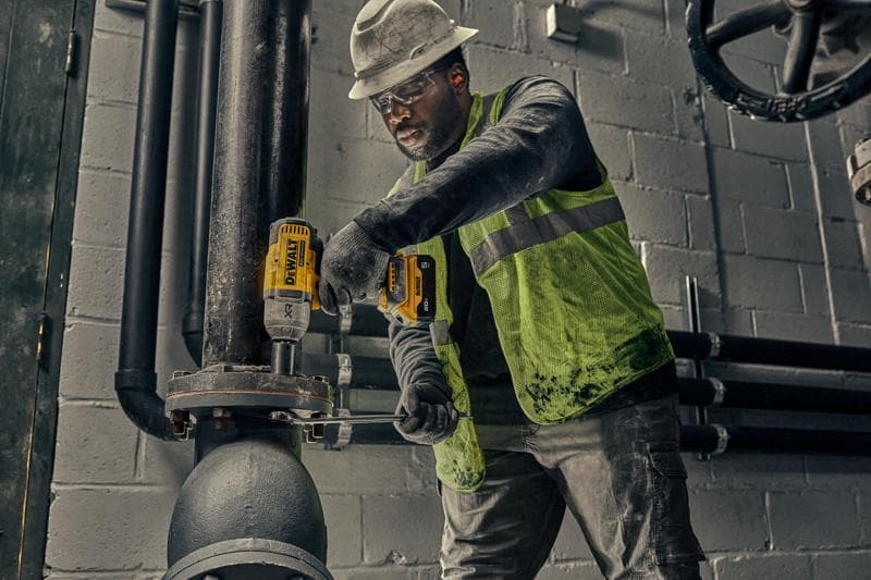 A worker wearing a hard hat and safety vest uses a yellow DEWALT power tool to secure bolts on a large industrial pipe inside a facility.