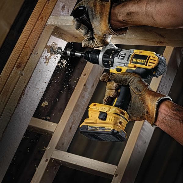 A person wearing brown work gloves uses a DEWALT cordless power drill to bore a hole in a wooden beam, with wood shavings flying from the drilling process.