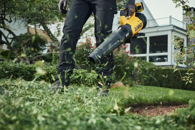 Person using a yellow and black DEWALT cordless leaf blower to clear grass clippings and leaves in a garden or yard.