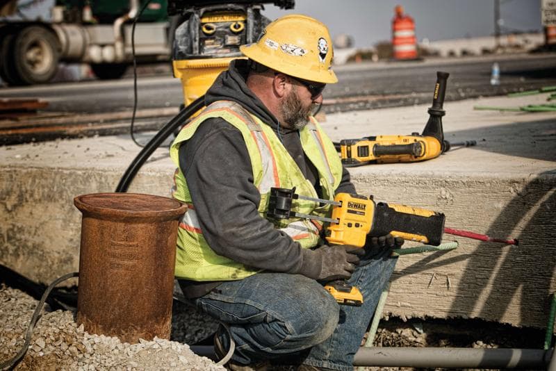 A construction worker wearing a yellow hard hat, safety vest, and gloves is using a DEWALT power tool to drill into concrete at an outdoor construction site. The worker's face is blurred for privacy. Other DEWALT tools and construction equipment are visible in the background.
