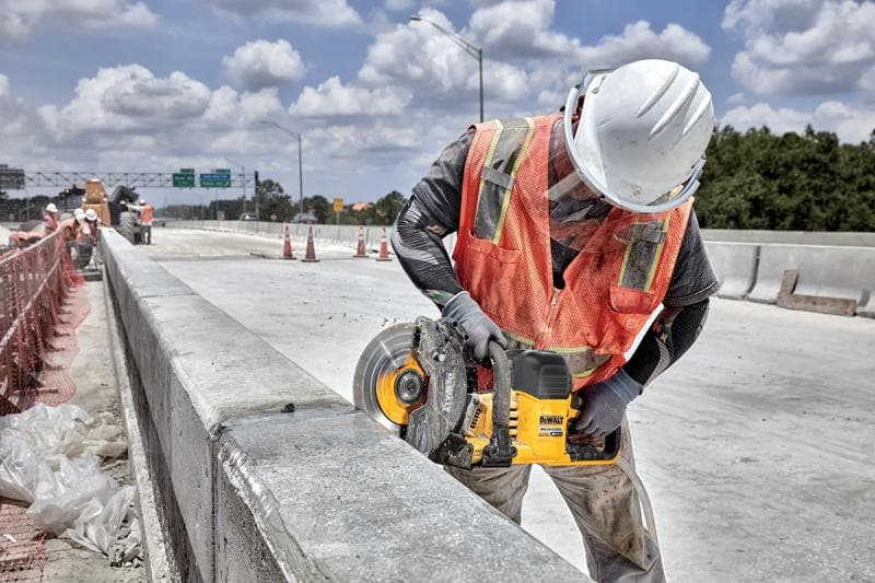 A construction worker wearing a white hard hat and orange safety vest is using a DEWALT cordless cut-off saw (model DCS692B) to cut through a concrete barrier on a road construction site.