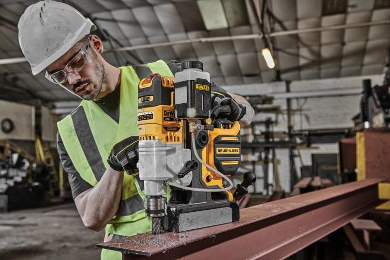 A person wearing a safety helmet and high-visibility vest is operating a DEWALT DCD1624 brushless magnetic drill on a steel beam inside an industrial workshop.