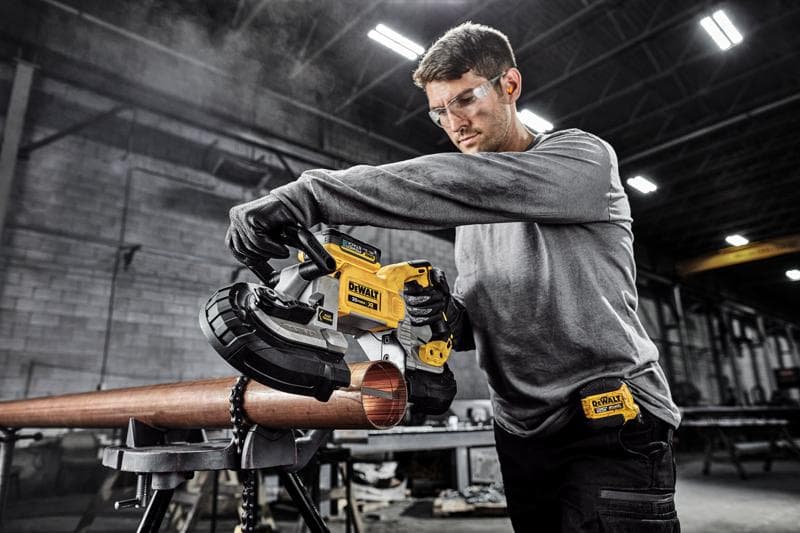 A person using a DEWALT DCS374B portable band saw to cut a metal pipe in an industrial workshop. The person's face is blurred for privacy.
