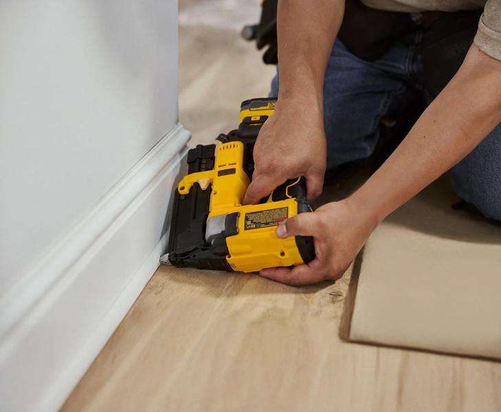 A person kneeling on the floor uses a yellow DEWALT cordless nailer to secure base molding to the wall in a home interior setting.