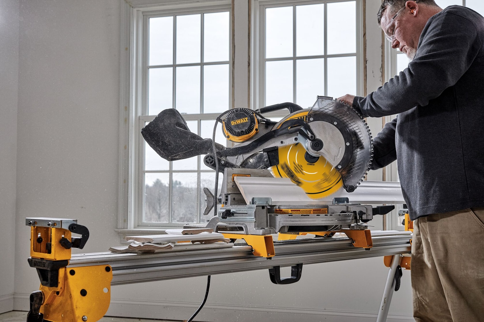A person uses a DEWALT miter saw to cut wood trim in a bright room, with the saw placed on a yellow and silver stand. The scene shows windows in the background and woodworking in progress.