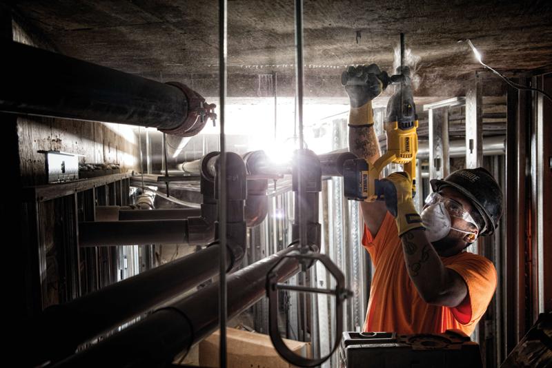 A person wearing safety gear uses a yellow DEWALT power tool to drill into a concrete ceiling in an industrial setting with pipes and metal structures.