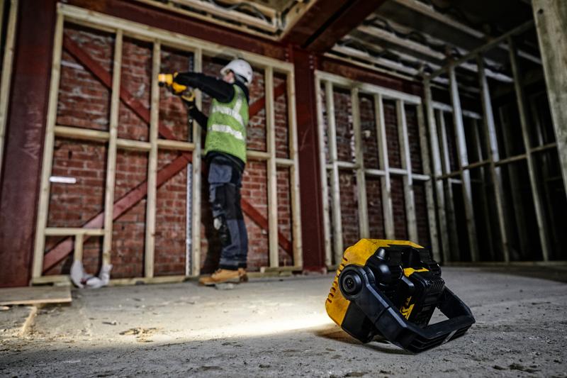 DEWALT DCL077 work light on floor in a construction site, worker in background using tools near wooden framing.