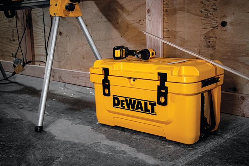 A yellow DEWALT toolbox placed on a concrete floor, with a power drill resting on top. The toolbox is situated near metal legs and a wooden wall in a workshop setting.