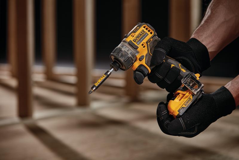 A close-up view of a person wearing black gloves holding a yellow DEWALT cordless drill driver with a bit attached, shown in a construction setting with wooden beams in the background.
