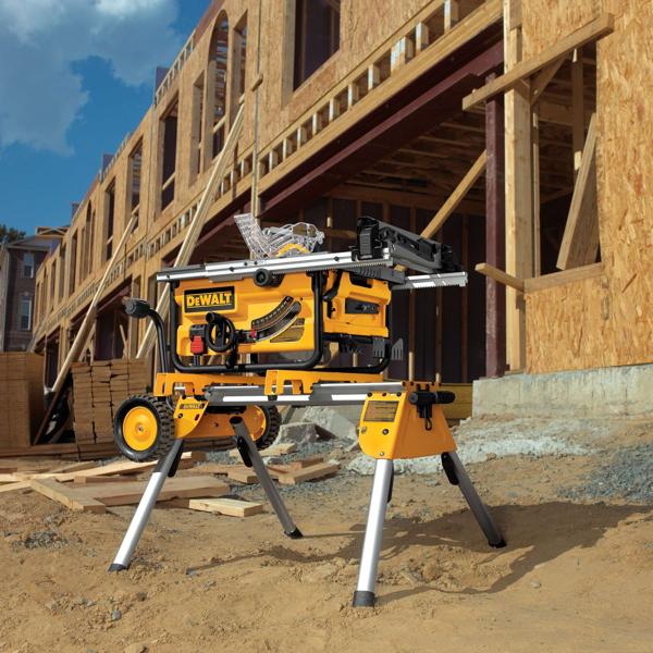 A DEWALT portable table saw with stand is shown on a construction site, with a partially constructed wooden building in the background. The saw and its stand are set on sandy ground, with building materials such as wooden planks stacked nearby.