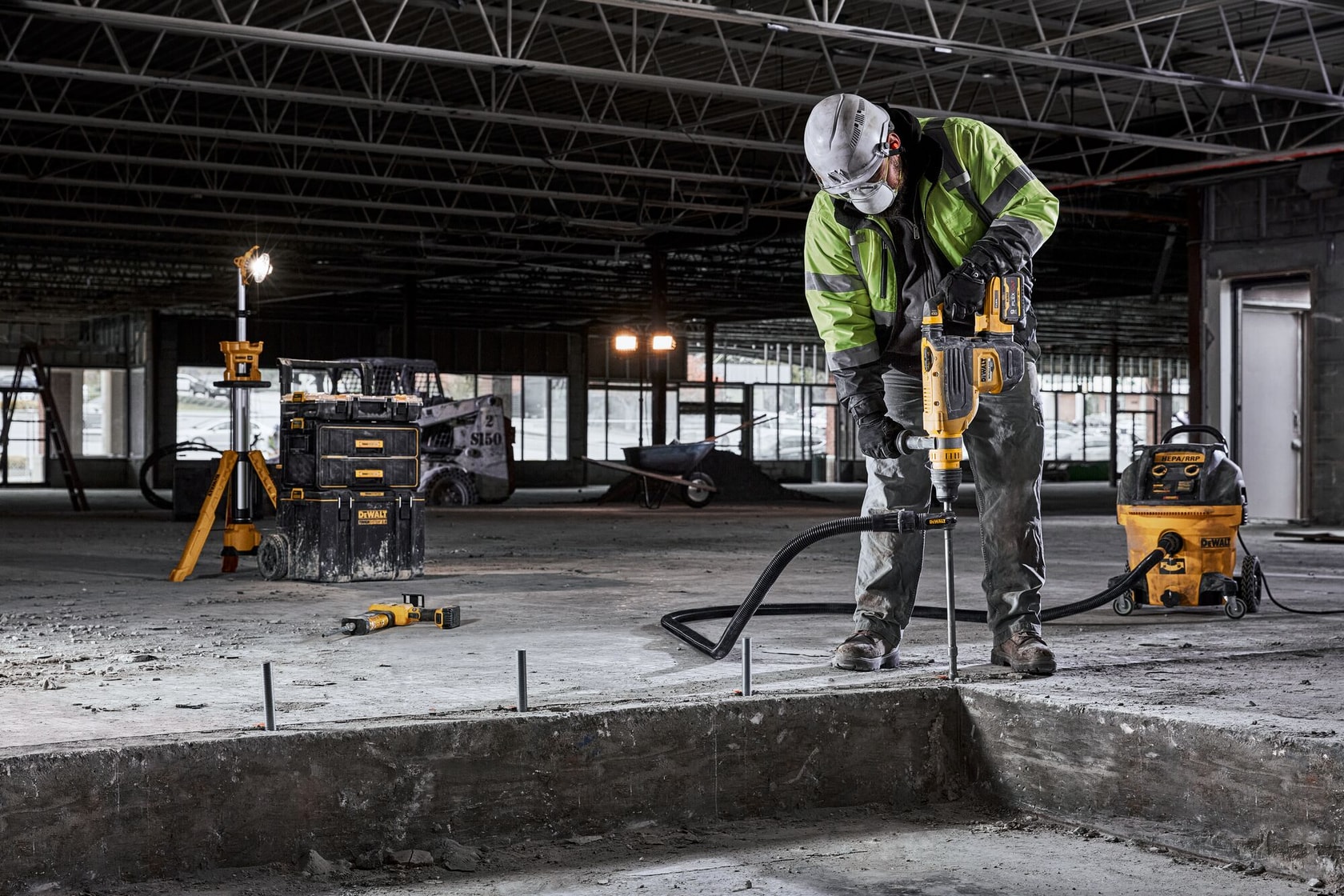 A construction worker wearing safety gear uses a DEWALT power tool to drill into concrete inside an unfinished building. Various DEWALT tools and equipment are visible around the work area.