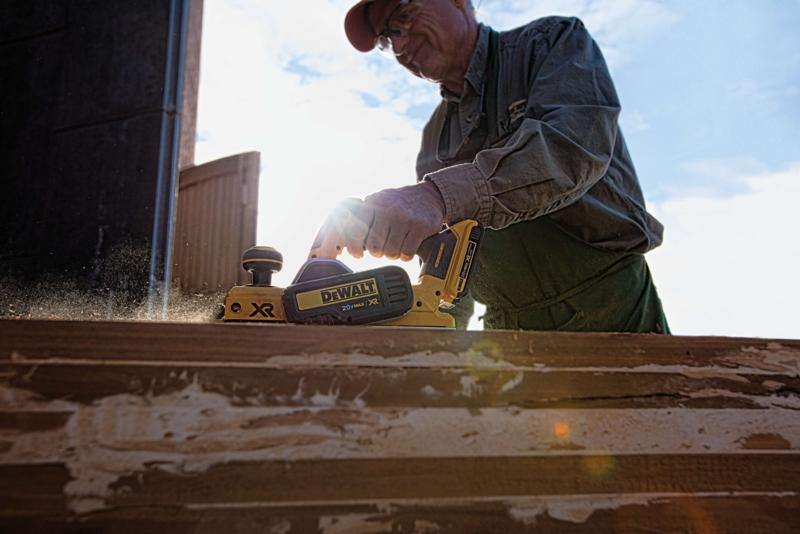 A person using a DEWALT DCP580B 20V cordless planer on a wooden surface outdoors, with wood shavings flying as the planer operates.